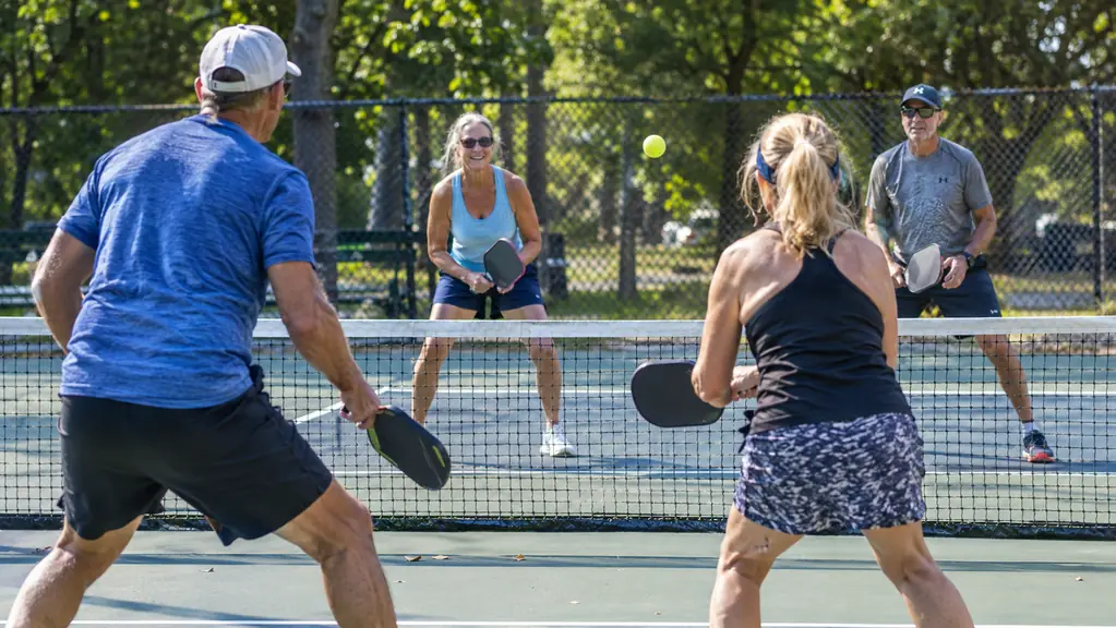 Recreational doubles players on a sunny park court, one player ready at the kitchen line, trees and a fence behind.