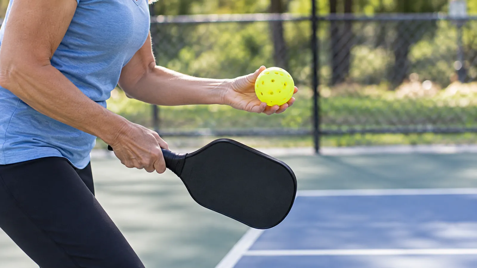 A player's hand holds a pickleball at the service line while the other hand holds a paddle ready to serve