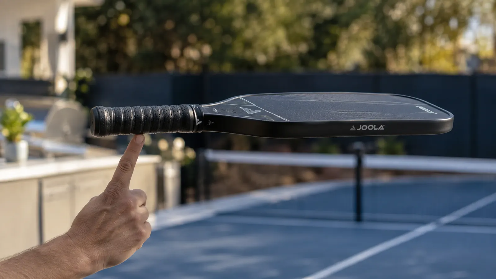 A pickleball paddle balanced on a player's fingertip at the grip, the kitchen line and court blurred in the background.