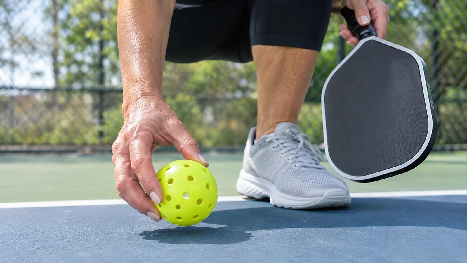 A hand picking up a pickleball from the court surface, paddle resting on the other knee
