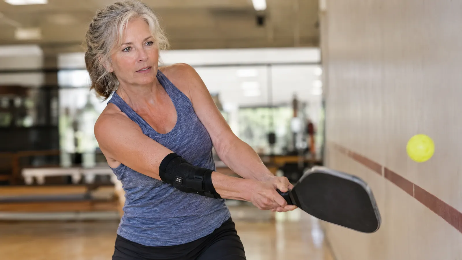A woman in her sixties practicing a two-handed backhand against a wall, paddle gripped loosely, brace on her forearm