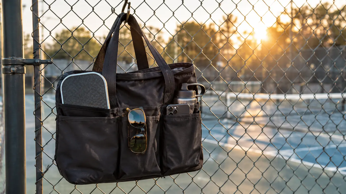 Large court bag hanging from a fence hook on a chain-link fence with a paddle and sunglasses visible in separate pockets.