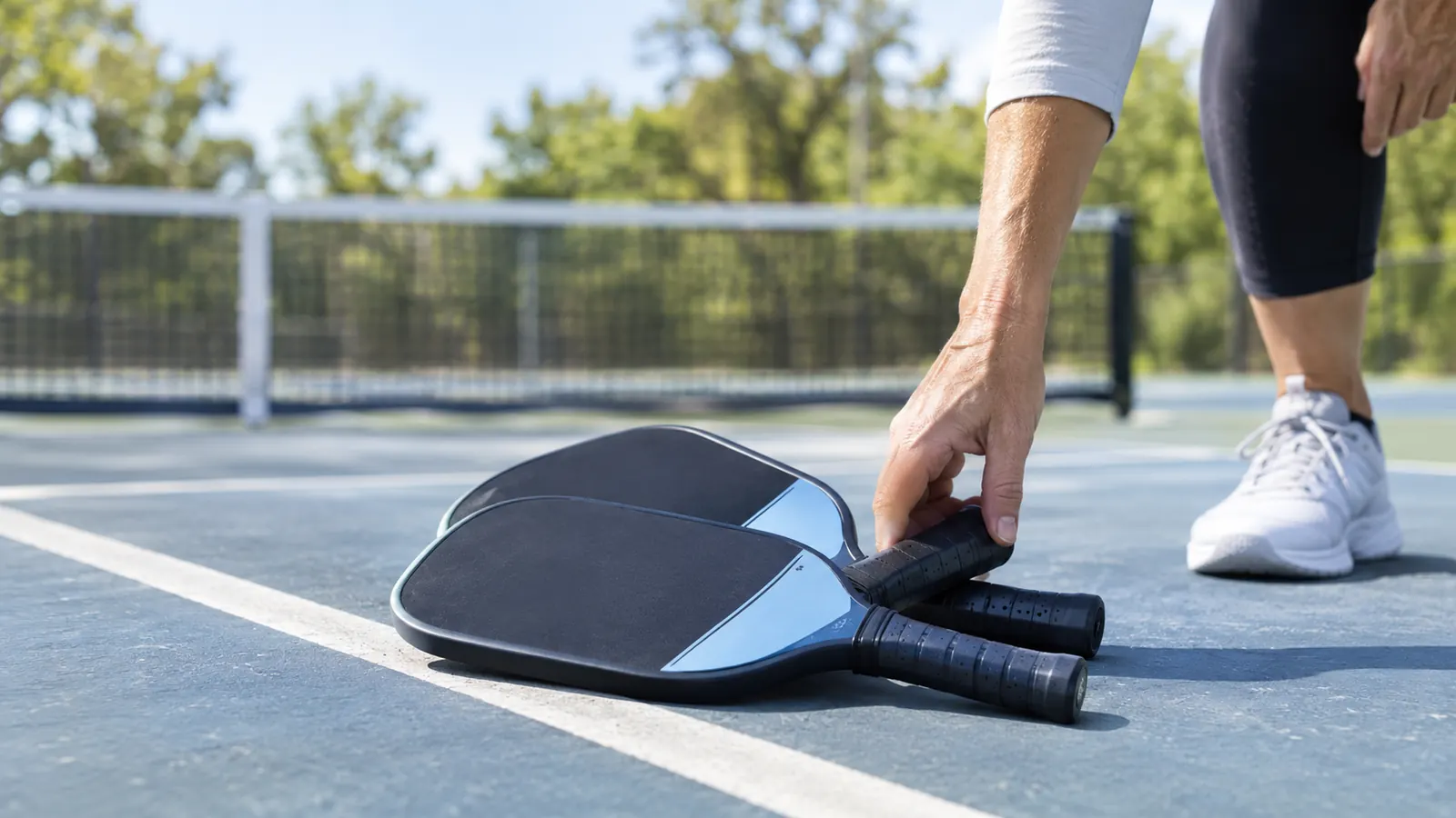 A hand picks up one paddle from a two-paddle set resting on the ground beside a pickleball court.