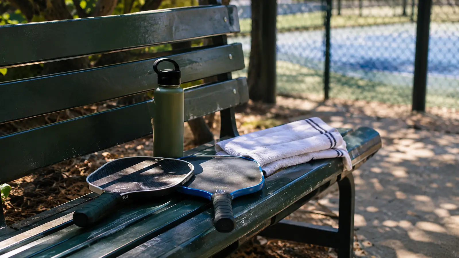 Two pickleball paddles and a towel on a bench in the shade after a morning of doubles