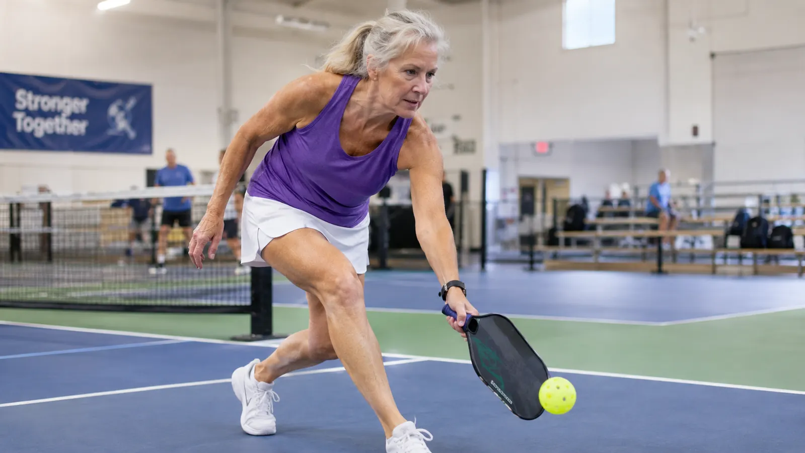 A woman in her sixties playing pickleball at the kitchen line, reaching for a low shot on an indoor court
